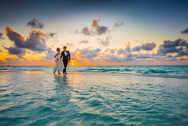 Žena v svadobných šatách a muž v smokingu kráčajú po pláži.jpg